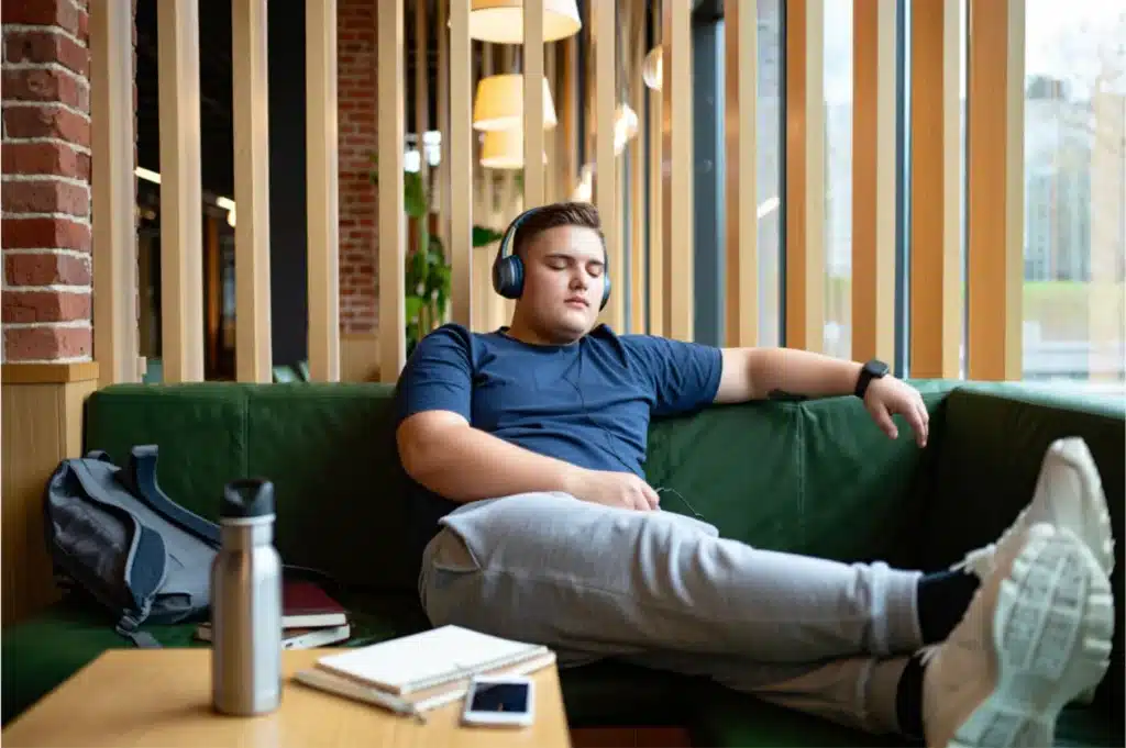A student reclines on a green couch in a quiet study space, eyes closed and wearing headphones, with a notebook and water bottle nearby. The scene illustrates using on-demand hypnotherapy recordings to reduce anxiety and mentally prepare for collaborative study sessions.